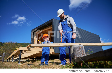 Father with toddler son building wooden frame house. Male builders hammering nail into plank on construction site, wearing helmet and blue overalls on sunny day. Carpentry and family concept. Father with toddler son building wooden frame house. Male builders hammering nail into plank on construction site, wearing helmet and blue overalls on sunny day. Carpentry and family concept. 90282756