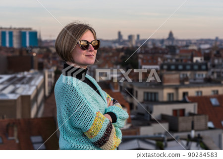 Portrait of an attractive thirty year old white woman with sunglasses posing on her rooftop with the Brussels skyline during sunset in the background 90284583