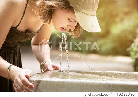 Teenage girl drinking water from an outdoor water fountain 90285386