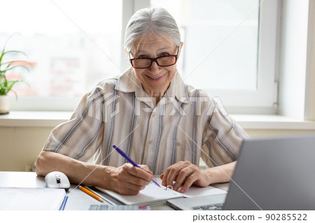 Friendly smiling caucasian senior woman in eyeglasses sits at the laptop with a pen in hand at home, selective focus. Friendly smiling caucasian senior woman in eyeglasses sits at the laptop with a pen in hand at home, selective focus. 90285522