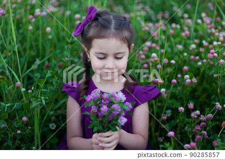 A beautiful little girl with long hair is smiling. A girl in a purple dress and a bouquet of clover in a blooming field 90285857