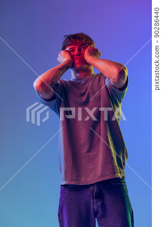 Young red-headed man, student grimacing isolated over blue studio background in neon light. Concept of youth, fashion, emotions, facial expression 90286440
