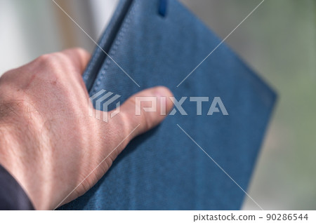 A man's hand holds a closed diary. Blue hardcover notebook. Close-up. Selective focus. A man's hand holds a closed diary. Blue hardcover notebook. Close-up. Selective focus. 90286544