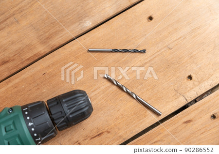 An electric drill and two drills lie against the wooden surface. The hand-held power tool is turning the boards. Selective focus. No people. 90286552
