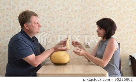 A happy married couple, a man and a woman, are having a fun conversation sitting at a table and holding a fragrant melon in their hands 90286962
