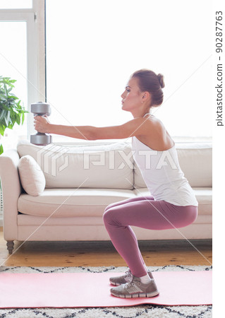 Young adult woman doing fitness exercises with gray dumbbells at her home in living room 90287763