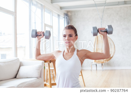 Athletic woman in sportswear doing fitness exercises with dumbbells at home. 90287764