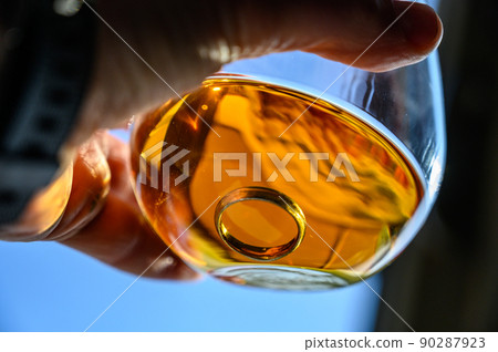 Caucasian hand holding a glass of amber colored liquid with a wedding ring at the bottom 90287923