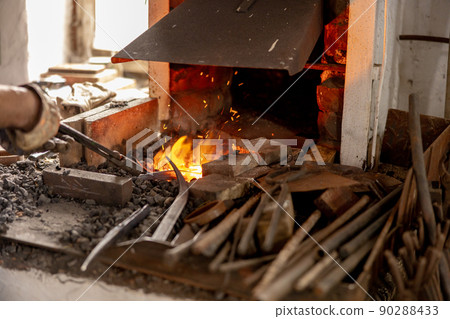 The blacksmith in production process of metal products, handmade in the forge. Craftsman heats metal in the fire. Metal industry, old profession. The blacksmith in production process of metal products, handmade in the forge. Craftsman heats metal in the fire. Metal industry, old profession. 90288433