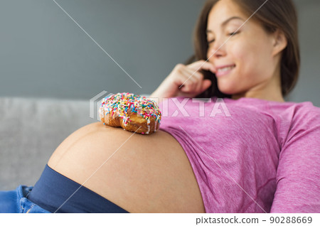 Unhealthy food during pregnancy. Sugar temptation craving Asian pregnant woman trying to resist eating a sweet donut on her baby belly. Funny eating cravings concept 90288669