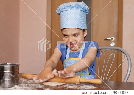 5 year old girl in blue dress and dressed as chef in kitchen laughs and rolling out dough 90288783
