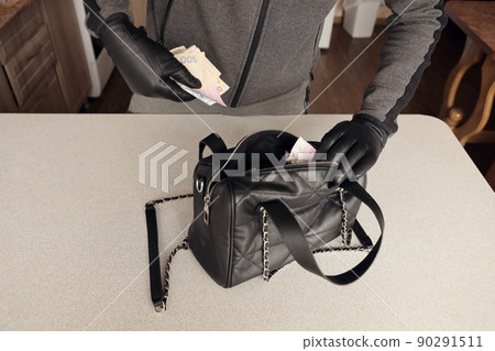 Robber in black outfit and gloves see in opened stolen women bag. The thief takes out the ukrainian money from a womans handbag in kitchen 90291511