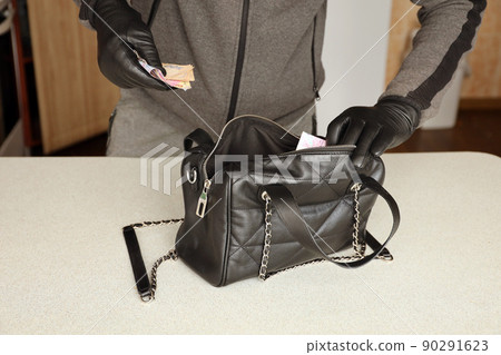 Robber in black outfit and gloves see in opened stolen women bag. The thief takes out the ukrainian money from a womans handbag in kitchen 90291623