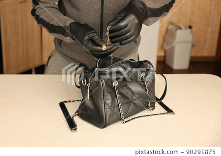 Robber in black outfit and gloves see in opened stolen women bag. The thief takes out golden necklace from a womans handbag in kitchen Robber in black outfit and gloves see in opened stolen women bag. The thief takes out golden necklace from a womans handbag in kitchen 90291875