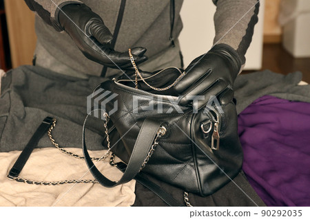 Robber in black outfit and gloves see in opened stolen women bag. The thief takes out golden necklace from a womans handbag in kitchen Robber in black outfit and gloves see in opened stolen women bag. The thief takes out golden necklace from a womans handbag in kitchen 90292035
