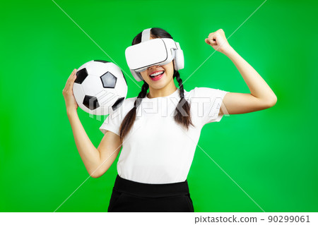 Cheerful asian girl football fan in vr glasses holding a soccer ball isolated green wall background, football sport and Metaverse Concepts 90299061