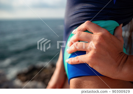 A young beautiful tanned woman with long hair in blue bikini stands at rock near blue green clear ocean sea water. Woman relax on sunset at sea, enjoy holidays and weekend vacation in summer time. 90300929
