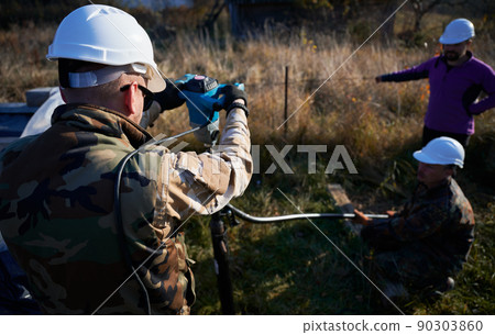 Male workers building pile foundation for wooden frame house. Men builders in white safety helmets drilling piles into the ground.. 90303860