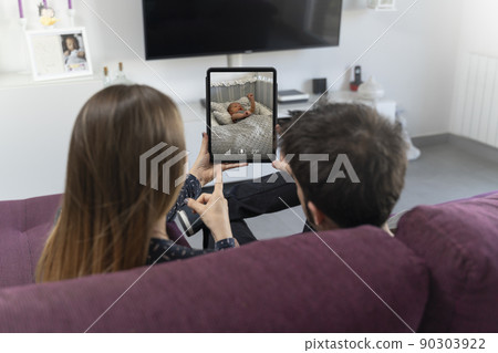 Two parents sitting on the sofa and monitoring their baby on the tablet 90303922