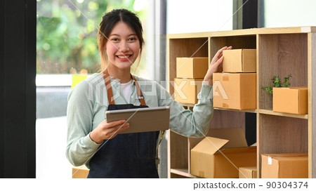 Confident female small business entrepreneur in apron holding digital tablet and working at warehouse for online seller 90304374