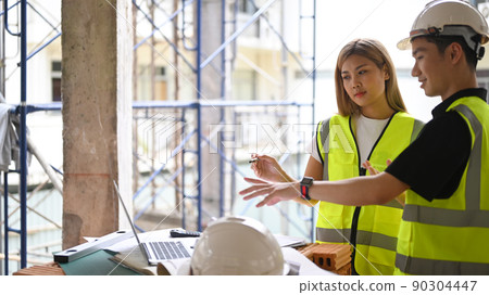 Two young engineers in helmets discussing construction plan at construction site. Industry, Engineer, construction concept Two young engineers in helmets discussing construction plan at construction site. Industry, Engineer, construction concept 90304447