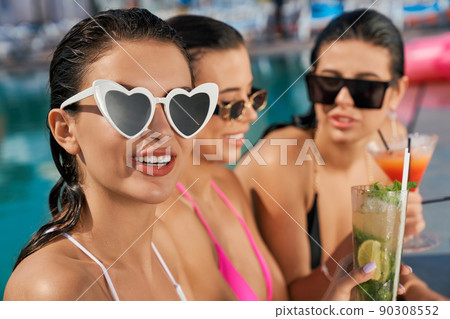 Close up of three pretty girl in swimsuits and sunglasses having vacation. Seductive young females standing in swimming pool, laughing, drinking cocktails. Concept of youth. 90308552