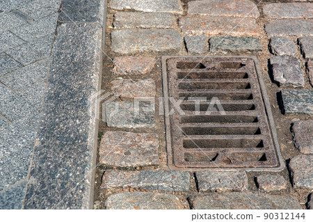 rusted rectangular grating manhole drainage system on the pavement road nearby to the gray granite curb and stone tile pedestrian pavement close-up on dry weather, nobody. rusted rectangular grating manhole drainage system on the pavement road nearby to the gray granite curb and stone tile pedestrian pavement close-up on dry weather, nobody. 90312144