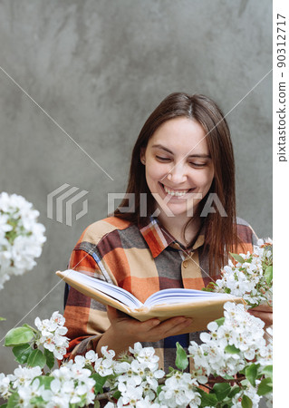 The girl reads a book wrapped in kraft paper and smiles sincerely against the background of a structured concrete wall. Defocused blurred Apple Flowers blossoms in the foreground. Spring concept The girl reads a book wrapped in kraft paper and smiles sincerely against the background of a structured concrete wall. Defocused blurred Apple Flowers blossoms in the foreground. Spring concept 90312717