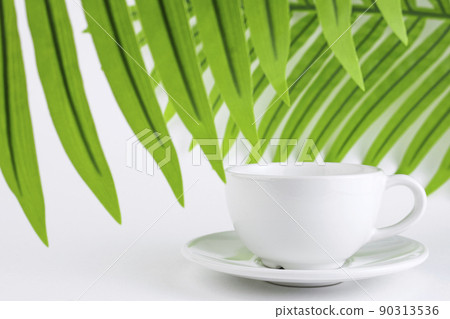 White mug and saucer on a white background, near palm leaves. A set of porcelain or ceramic dishes. Copy space 90313536