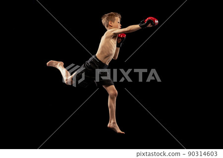 Studio shot of preschool boy, cute kid in sports shots and gloves training alone on black background. Sport, education, action, motion concept. 90314603