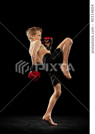 Studio shot of preschool boy, cute kid in sports shots and gloves training alone on black background. Sport, education, action, motion concept. 90314604