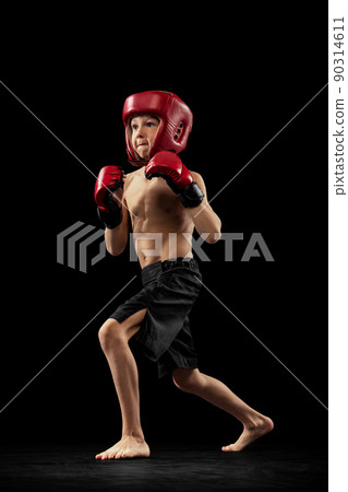 Studio shot of preschool boy, cute kid in sports shots and gloves training alone on black background. Sport, education, action, motion concept. 90314611