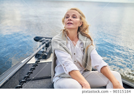 A blonde mature woman in white clothes sitting on a boat and looking dreamy 90315184