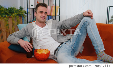 Man sitting on sofa eating chips and watching interesting TV serial, football game online at home Man sitting on sofa eating chips and watching interesting TV serial, football game online at home 90317114