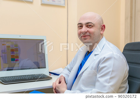 A male ophthalmologist at the workplace at the computer looks at the camera with a smile 90320172