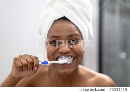 Portrait of african american mid adult woman in towel brushing teeth in bathroom 90335429
