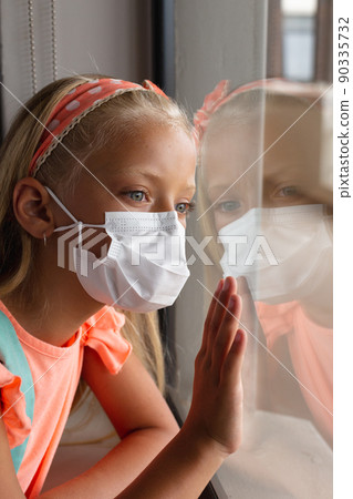 Close-up of caucasian elementary schoolgirl wearing mask while looking through window 90335732