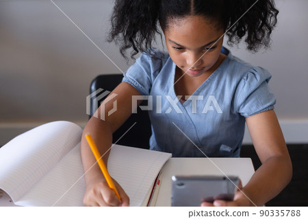 High angle view of biracial elementary schoolgirl using digital tablet while writing on book at desk 90335788
