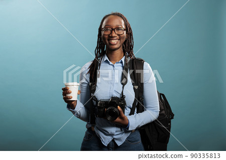 Happy positive photography entusiast having DSLR photo device and cup of coffee while standing on blue background. Young adult woman with photographing hobby smiling at camera. Happy positive photography entusiast having DSLR photo device and cup of coffee while standing on blue background. Young adult woman with photographing hobby smiling at camera. 90335813