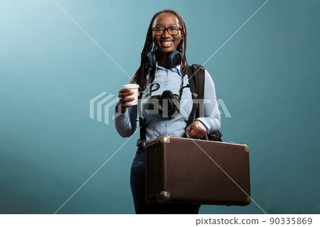 Happy traveler with holiday suitcase and DSLR camera smiling optimistic at camera while getting ready for vacation. Smiling tourist with photo device and wireless headphones waiting to go on journey. 90335869