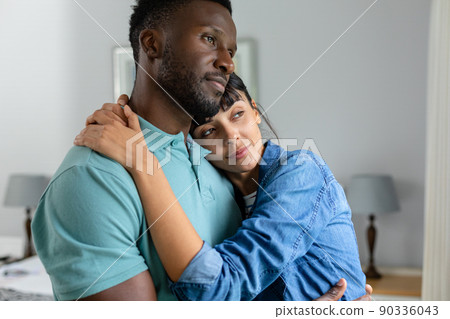 Contemplative multiracial young couple embracing while standing together at home Contemplative multiracial young couple embracing while standing together at home 90336043