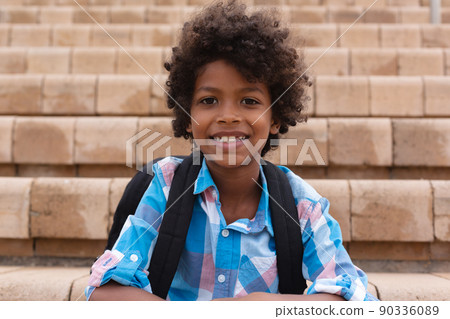 Portrait of smiling african american elementary boy with backpack sitting on school building steps Portrait of smiling african american elementary boy with backpack sitting on school building steps 90336089