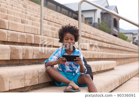 Smiling african american elementary schoolboy using smart phone while sitting on school steps Smiling african american elementary schoolboy using smart phone while sitting on school steps 90336091