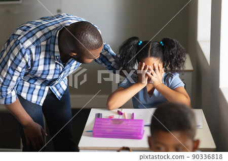 African american young male teacher consoling sad biracial elementary schoolgirl sitting at desk African american young male teacher consoling sad biracial elementary schoolgirl sitting at desk 90336818