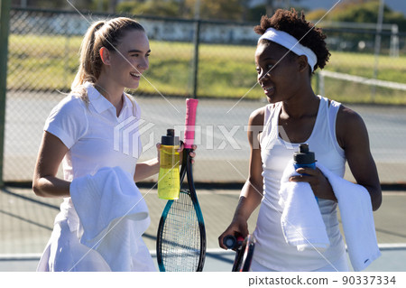 Smiling biracial female tennis players with bottles and towels talking during break at court Smiling biracial female tennis players with bottles and towels talking during break at court 90337334