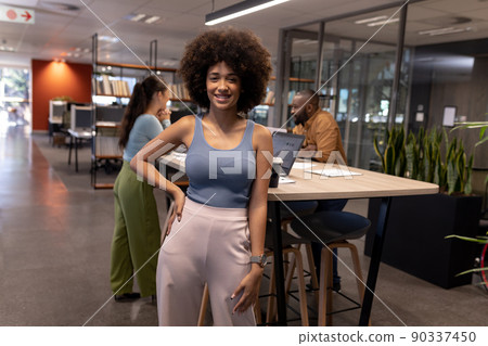Portrait of smiling young biracial businesswoman with afro hairstyle against colleagues at workplace 90337450