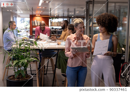 Smiling biracial businesswomen discussing together against colleagues working at modern workplace Smiling biracial businesswomen discussing together against colleagues working at modern workplace 90337461