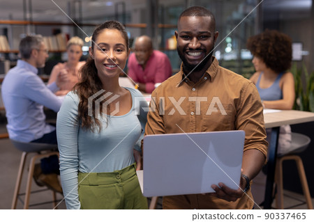 Portrait of smiling businessman and businesswoman with laptop against colleagues working at office Portrait of smiling businessman and businesswoman with laptop against colleagues working at office 90337465
