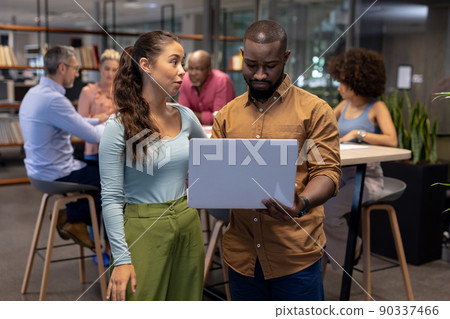Young biracial businessman and businesswoman discussing over laptop against colleagues at workplace Young biracial businessman and businesswoman discussing over laptop against colleagues at workplace 90337466