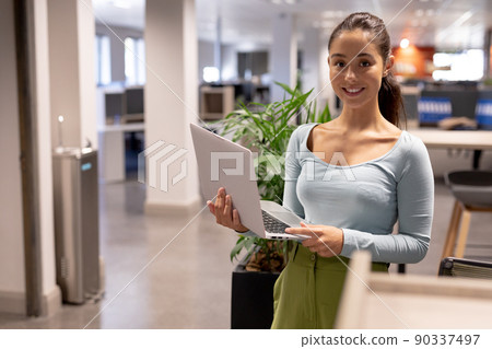 Portrait of smiling young female hispanic advisor holding laptop while working at modern workplace Portrait of smiling young female hispanic advisor holding laptop while working at modern workplace 90337497